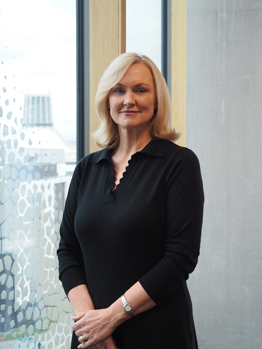 Joan is standing in front of a window with a view of Liverpool behind. She is smiling