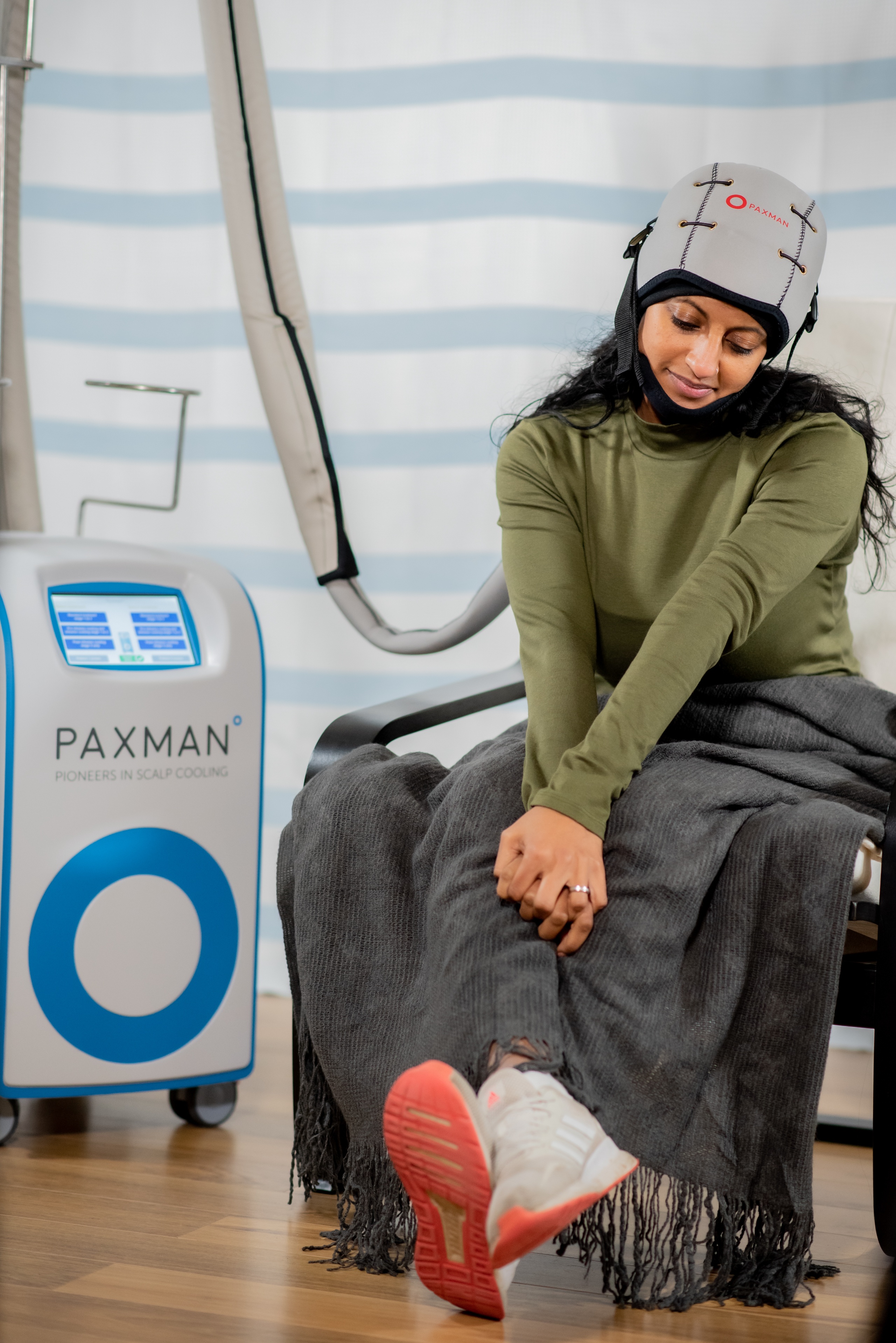 A young Asian women is wearing a cool cap for cancer treatment, The cap is thick grey fabric with a black chinstrap to keep it in position. It is connected to the Paxman scalp cooling machine by a long tube