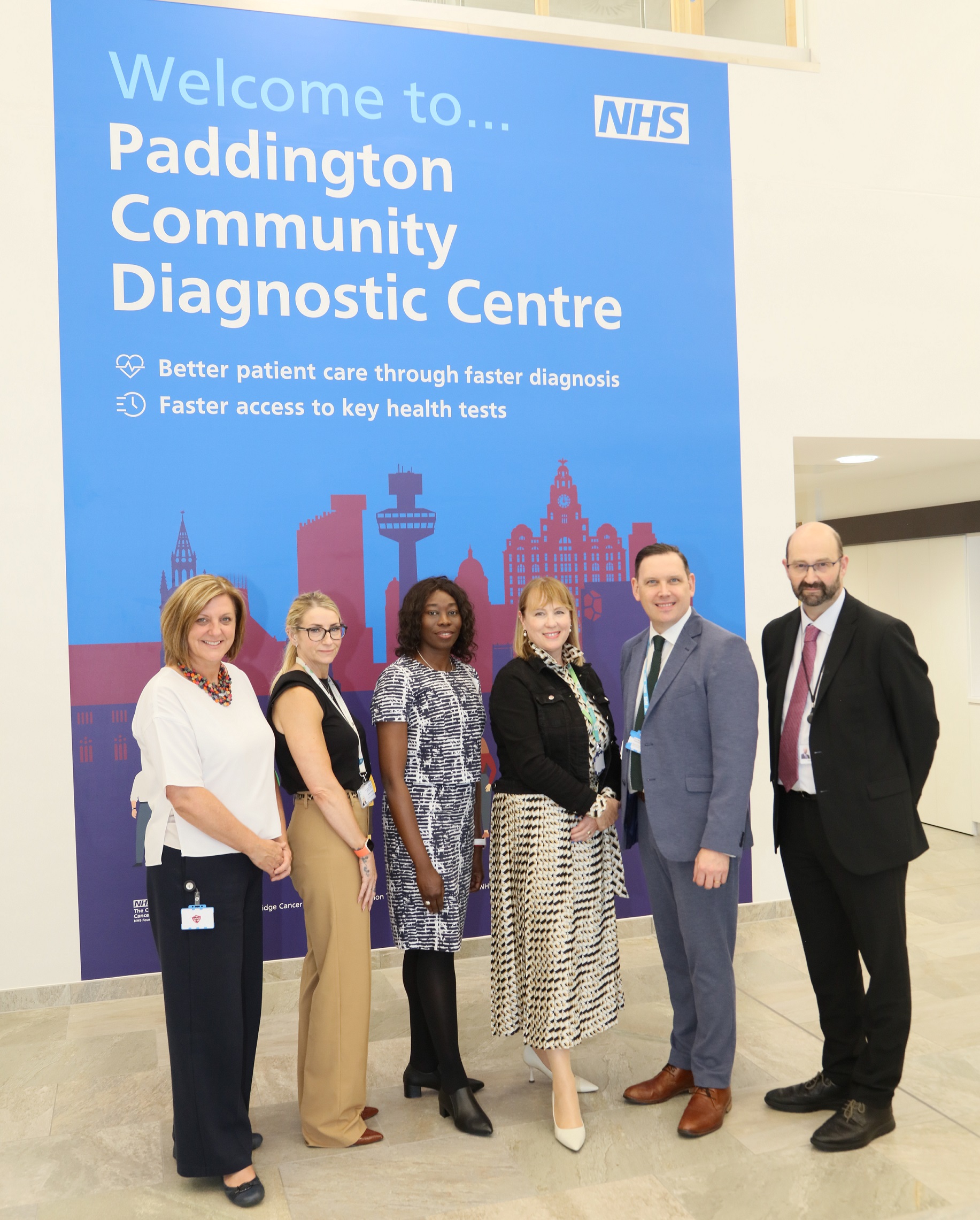 Picture of Chief Executive Liz Bishop with invited guests at the opening. They are pictured in front of a large graphic on the wall that says Paddington Community Diagnostic Centre and has artwork featuring landmark buildings across Liverpool and some people