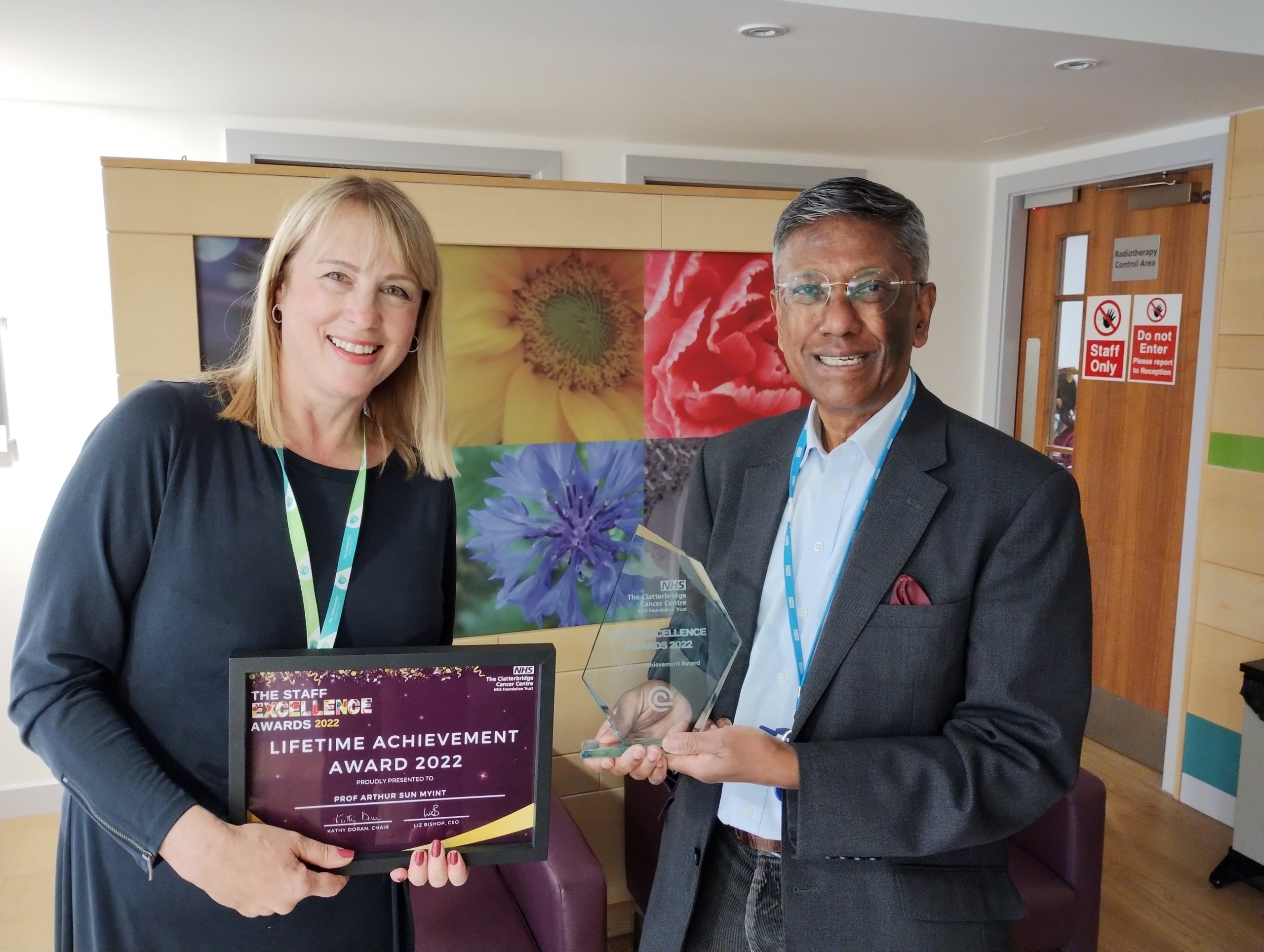 Chief Executive Liz Bishop is smiling and standing on the left holding a certificate that says 'Lifetime Achievement Award'. Prof Myint is on the right holding a glass awards trophy.