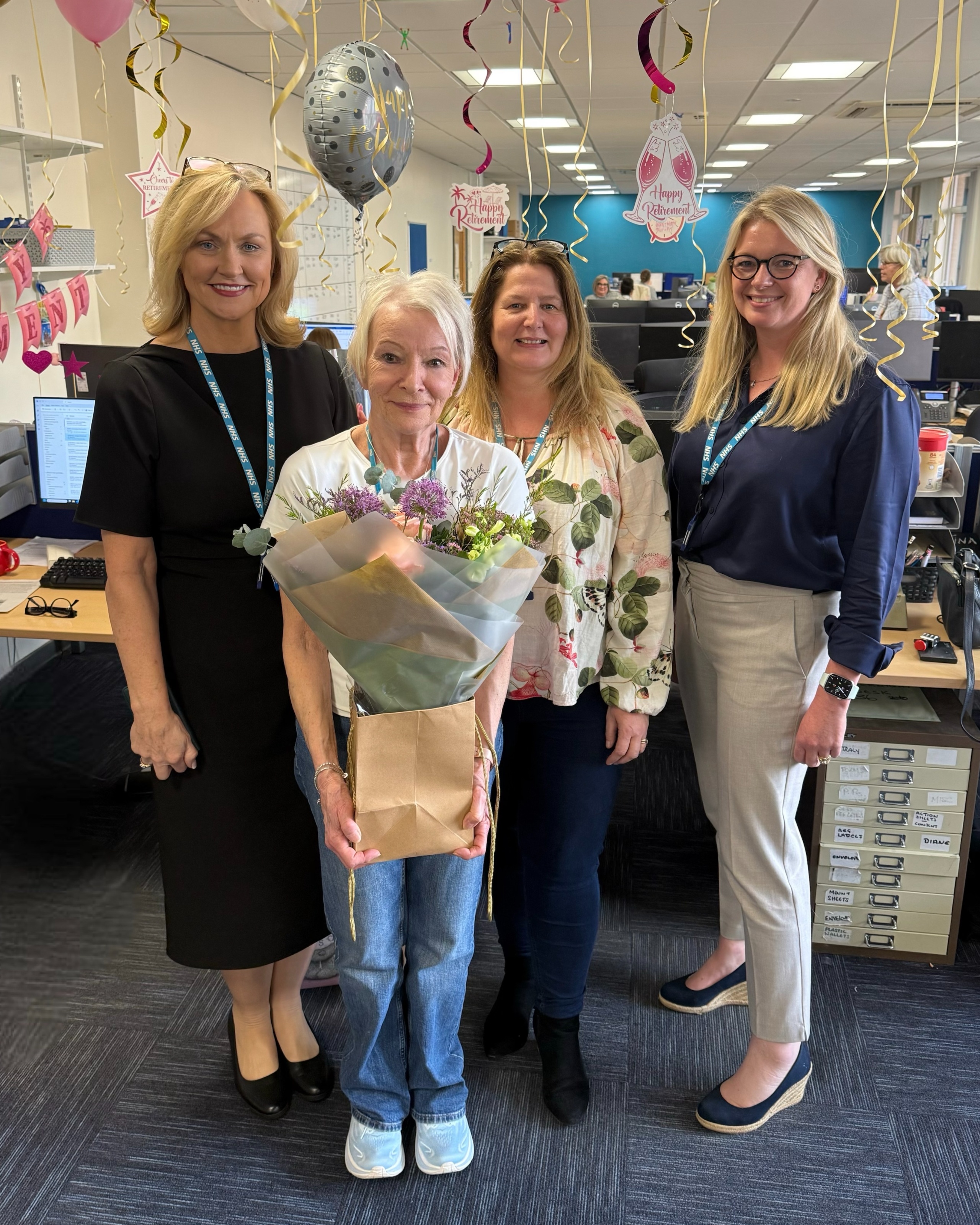 Cathy is standing in an office with 3 other women. She is holding a large bouquet of flowers. There are balloons behind them for a happy retirement. 