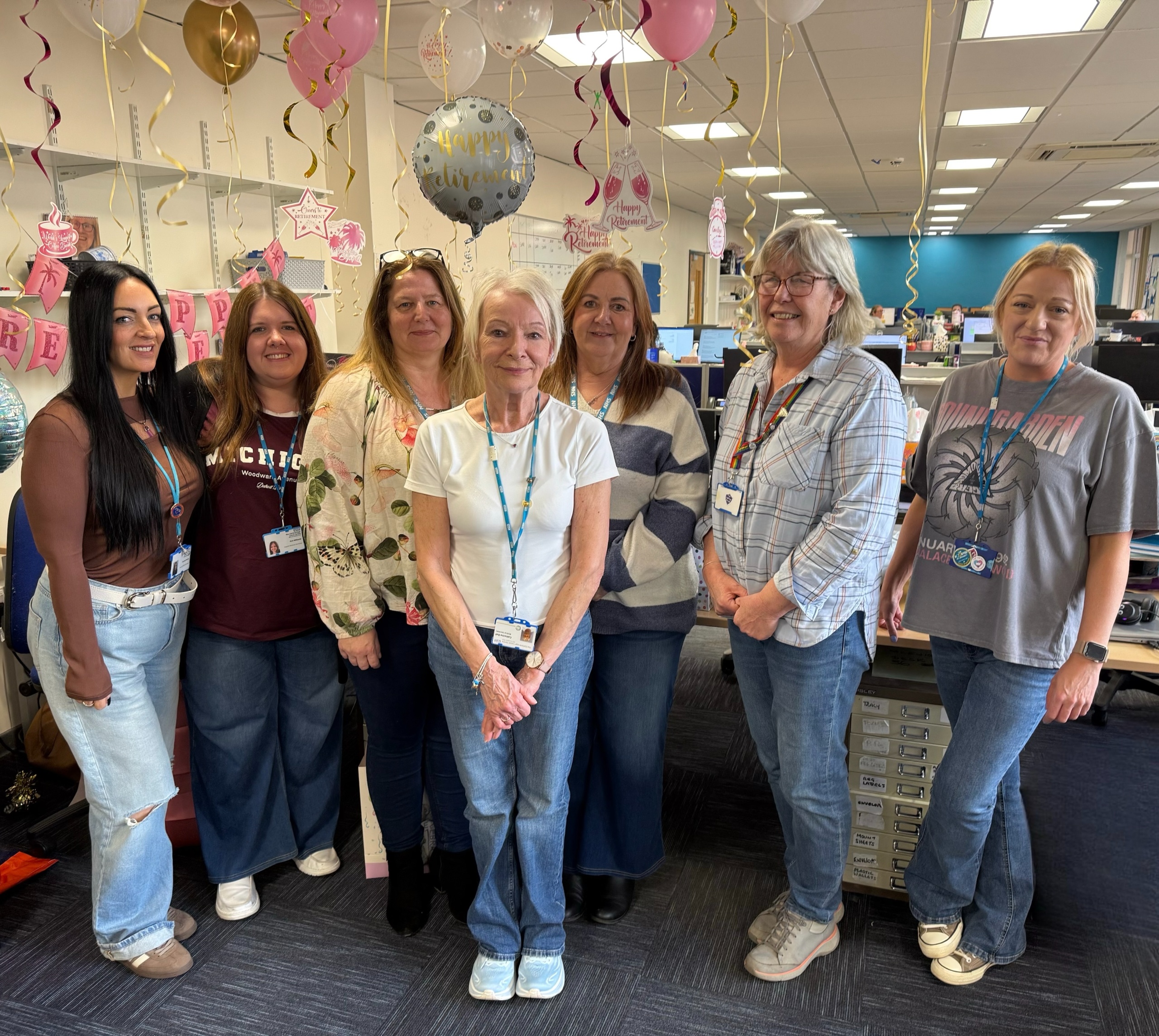 Cathy is standing with a group of her work colleagues. She is smiling. There are balloons behind them for a happy retirement.