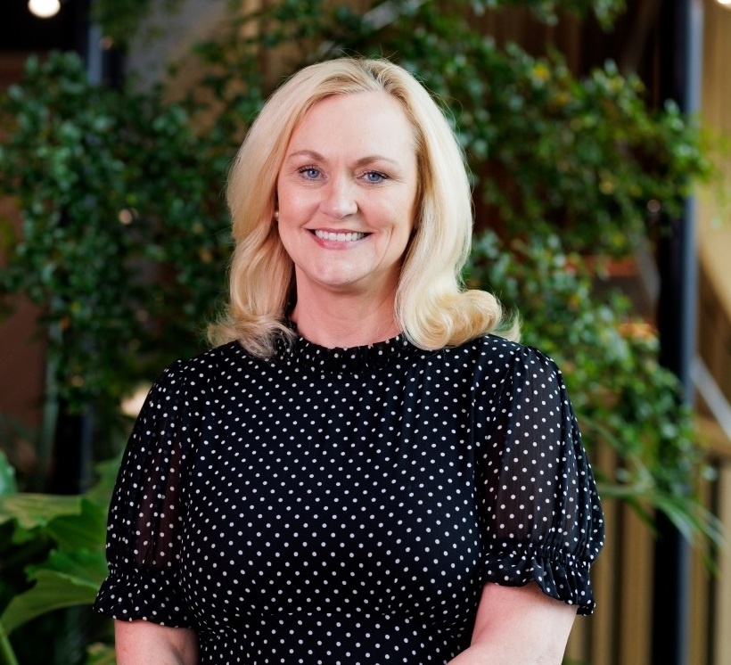 Joan has blonde hair and is wearing a black dress with white spots. She is smiling