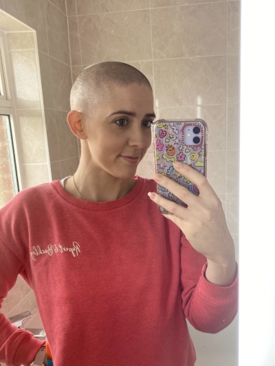 Vicky is taking a selfie in the mirror. She has shaved her head. She is wearing a pink jumper. 