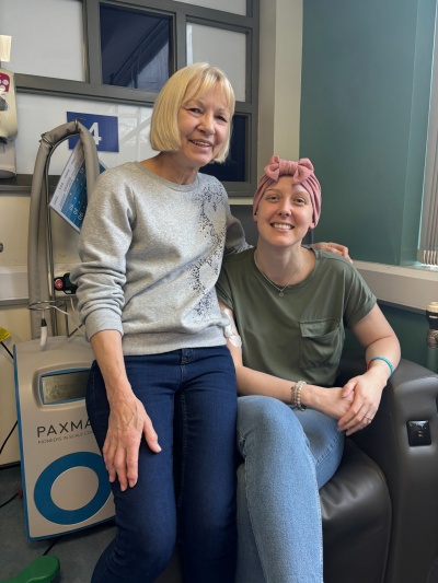Vicky (R) with her Mum Sue. Vicky is sitting in a chemotherapy chair. She is wearing a headscarf. Sue is sitting on the arm of the chair. 