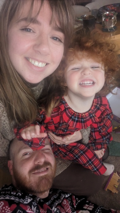Hannah smiles with her daughter and husband. Her daughter is 3 years old and had red curly hair. Her husband has a beard. They all smile for the camera. 