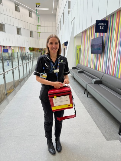Alison is standing in a bright hospital setting. She is holding a red square padded bag with a strap. It has a tag fitted to track where it is
