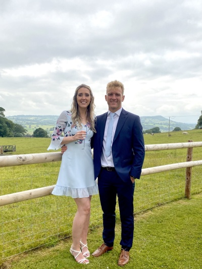 Vicky and Mick stand together in a field, holding champagne. She is dressed in a blue dress, he is in a suit. They are smiling 