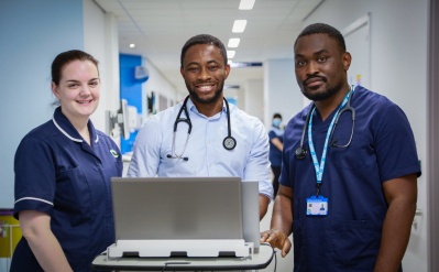 Two black doctors and white nurse on inpatient ward . They are at a mobile computer on wheels and are smiling