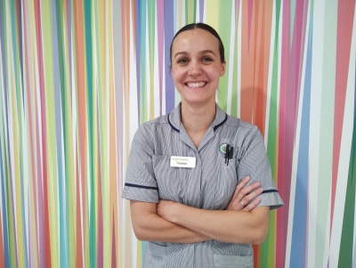 Chelcie has a blue and white striped nurses uniform. She is smiling and has her arms crossed. There is a colourful background behind her