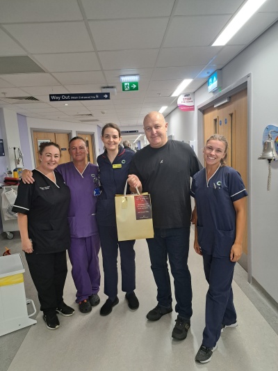 Will is pictured with hospital staff on the stem cell transplant unit. They are wearing clinical uniforms. He is wearing a jumper and holding a bag - one of the care packs he has donated for patients