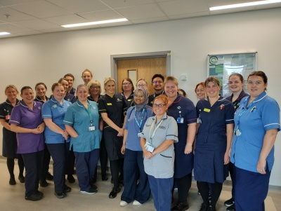 Large group of healthcare staff in different clinical uniforms on a hospital ward, smiling. 