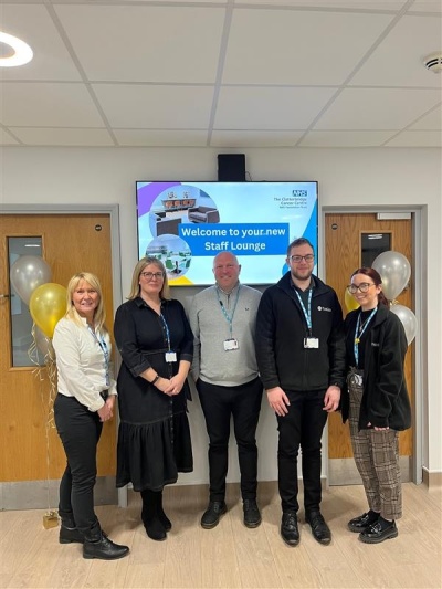 The Project Management Team stand infront of a TV which reads Welcom to your new Staff Lounge 