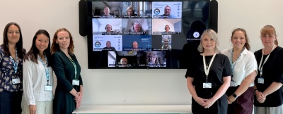 Group of people including some standing in front of a large screen and others on the screen in a Microsoft Teams meeting