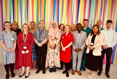 Group shot of consultants and other staff. They are a mix of men and women. Some are wearing scrubs or clinical uniforms. They are all smiling and are standing in front of a wall with brightly coloured stripes.